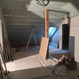 All-Bud Aleksander Sobczak - Poddasze w trakcie wykańczania płytami gipsowo-kartonowymi; widoczne łączenia szpachlowane, drabina, drewniany słup nośny oraz okno dachowe w skosie.