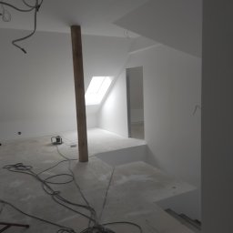 All-Bud Aleksander Sobczak - Poddasze w trakcie wykańczania, świeżo pomalowane na biało, widoczny drewniany słup podtrzymujący konstrukcję, okno dachowe i zwisające kable elektryczne.