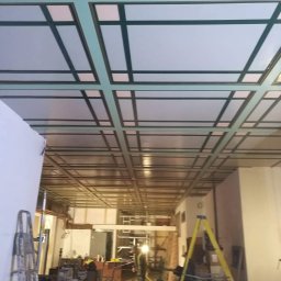 All-Bud Aleksander Sobczak - Sufit z widoczną konstrukcją nośną i częściowo zamontowanymi panelami, widoczne drabiny i sprzęt budowlany, w tle osoba w odblaskowej kamizelce.