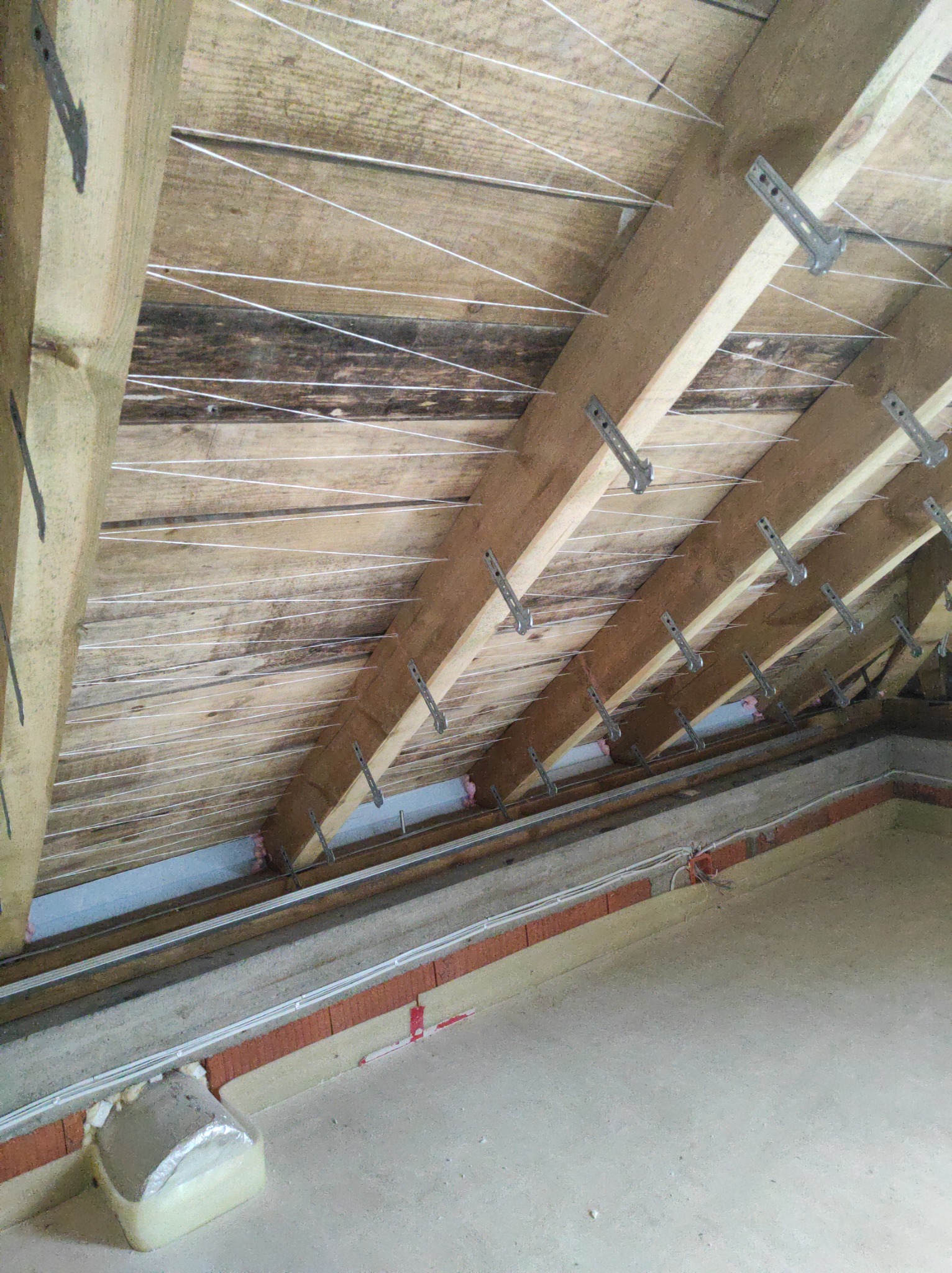 Drewniana konstrukcja dachu poddasza z zamocowanymi metalowymi uchwytami i rozciągniętymi białymi sznurkami, widoczne fragmenty izolacji przy ścianie kolankowej.