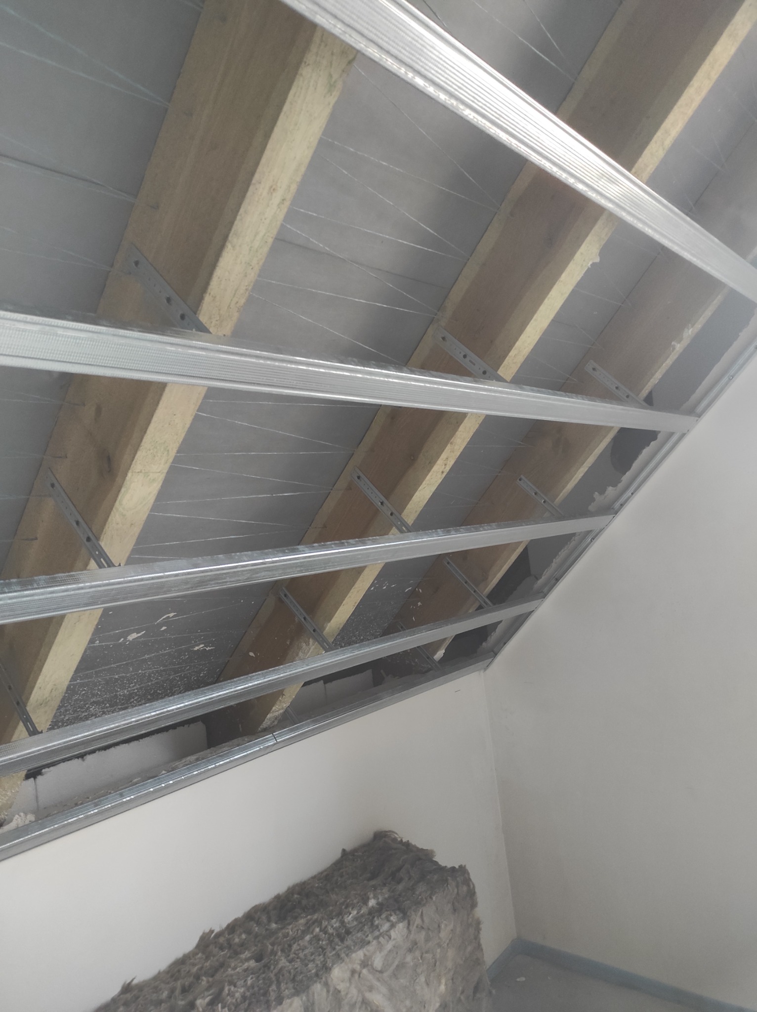 Konstrukcja metalowa sufitu podwieszanego z drewnianymi belkami, częściowo wypełniona materiałem izolacyjnym, widok z dołu.