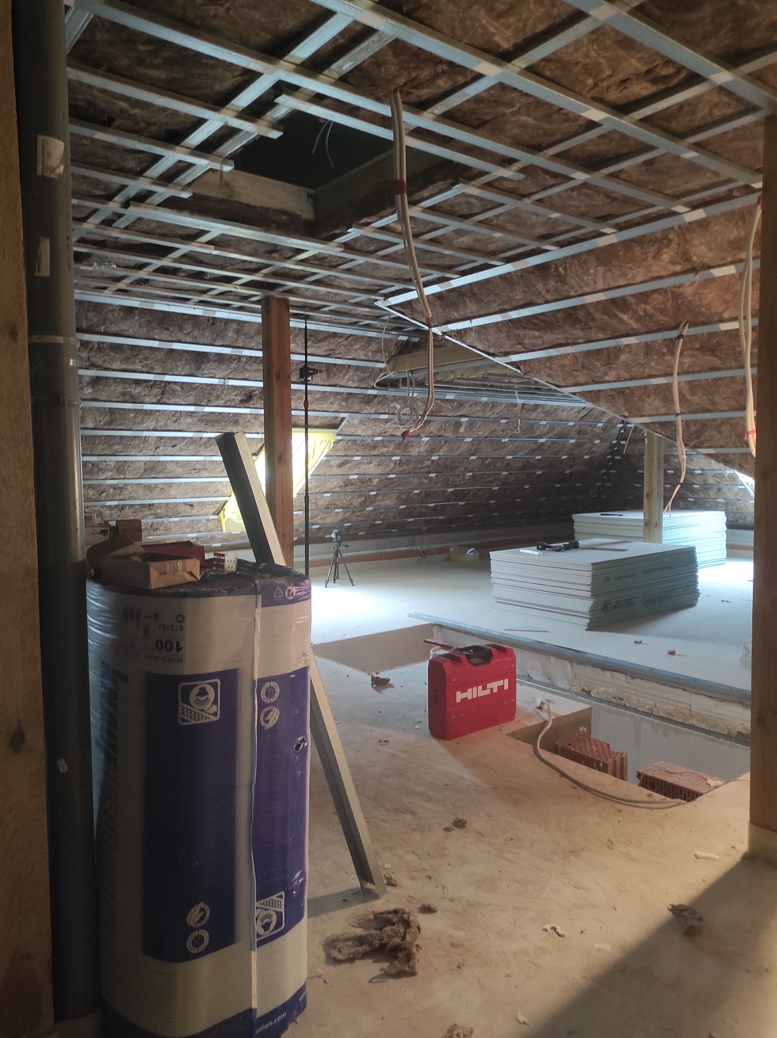 Poddasze w trakcie prac wykończeniowych: ocieplone skosy z widoczną konstrukcją metalową, rolka wełny mineralnej w folii, czerwona skrzynka narzędziowa Hilti, stos płyt gipsowo-kartonowych.