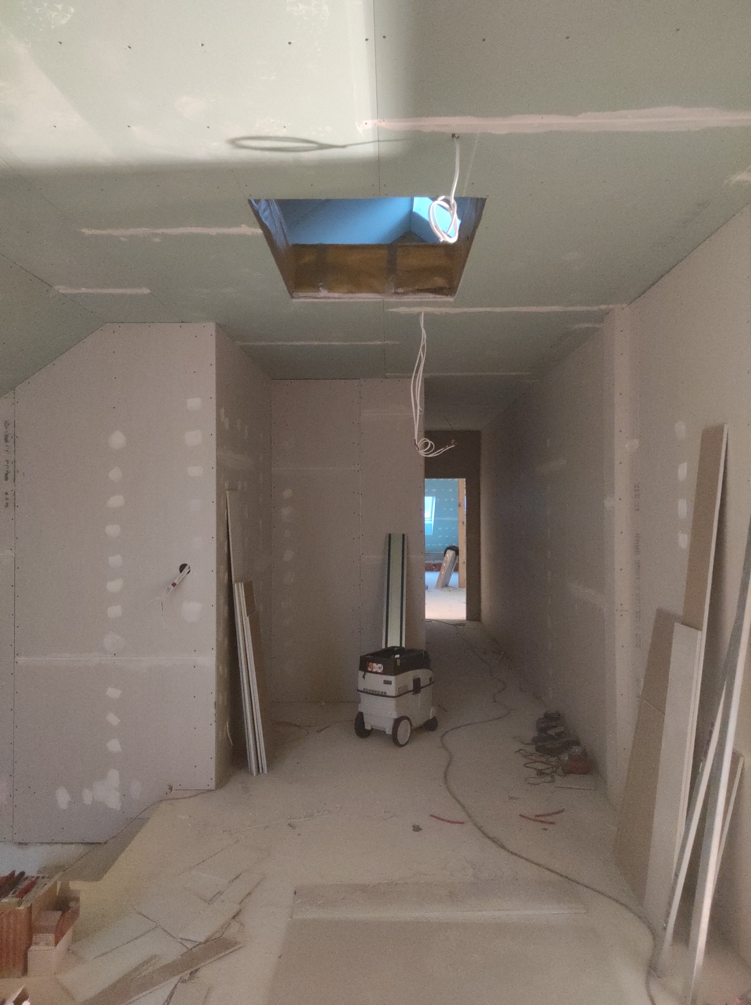 Korytarz w trakcie wykańczania: surowe ściany z płyt kartonowo-gipsowych, widoczne łączenia wymagające szpachlowania, otwór w suficie z wystającymi przewodami, odkurzacz przemysłowy i narzędzia...