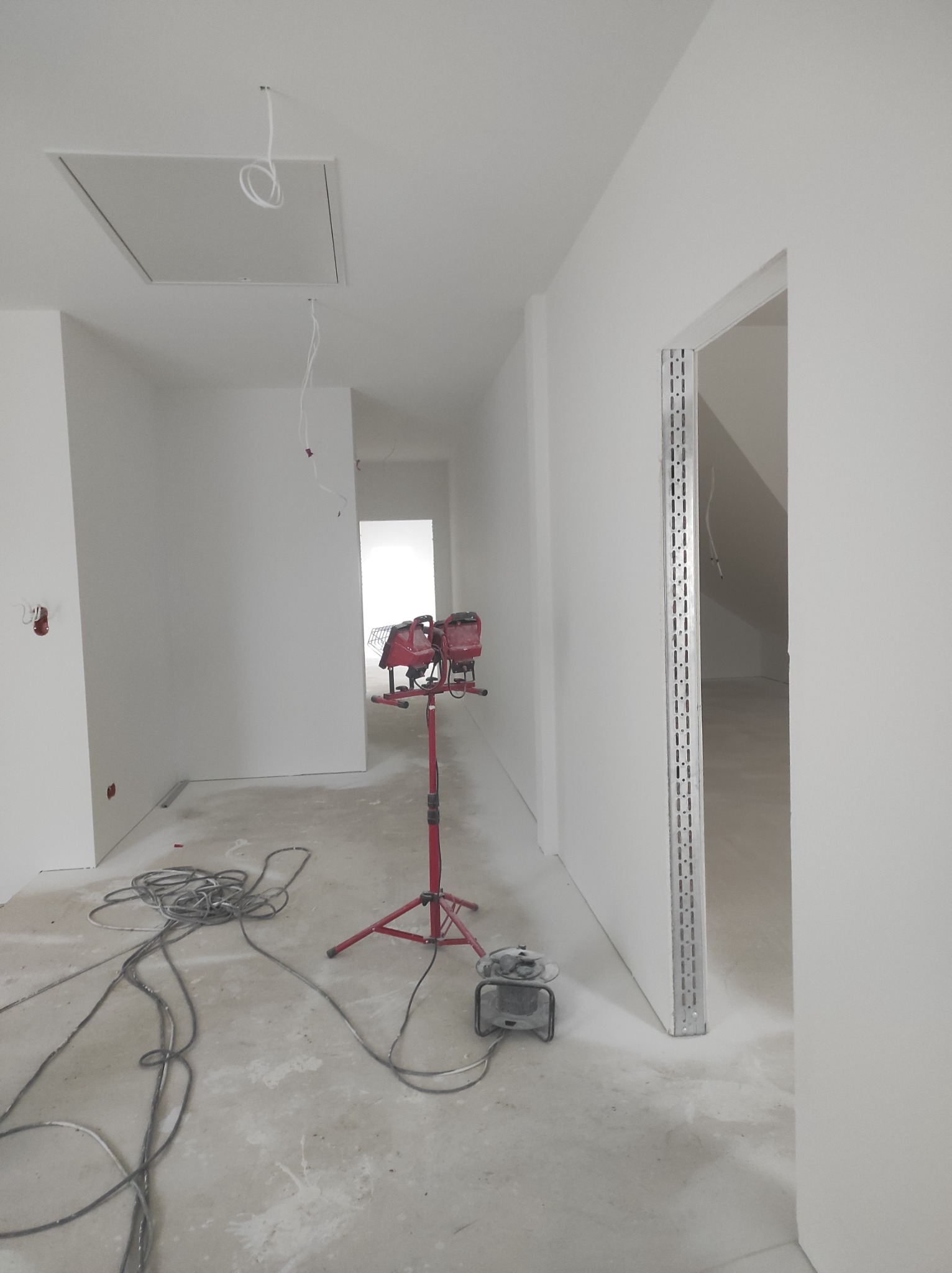 Wnętrze w trakcie wykańczania: białe ściany po szpachlowaniu i malowaniu, widoczne kable, lampa budowlana na trójnogu, szlifierka do gipsu oraz metalowe profile ościeżnic.