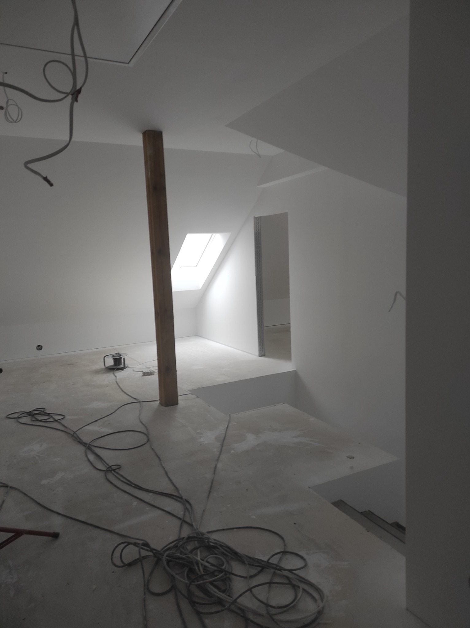 Poddasze w trakcie wykańczania, świeżo pomalowane na biało, widoczny drewniany słup podtrzymujący konstrukcję, okno dachowe i zwisające kable elektryczne.