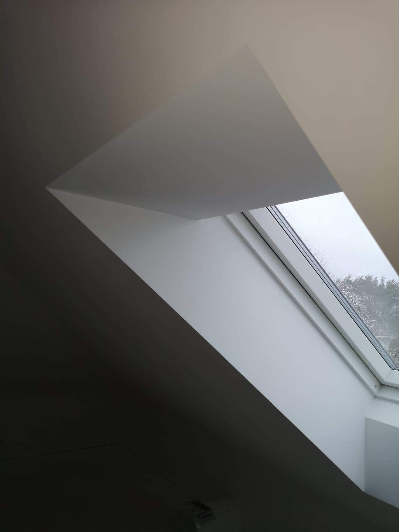 Wnętrze poddasza z oknem dachowym, widok na zewnątrz z kroplami deszczu na szybie, świeżo pomalowane ściany w odcieniach bieli i szarości.