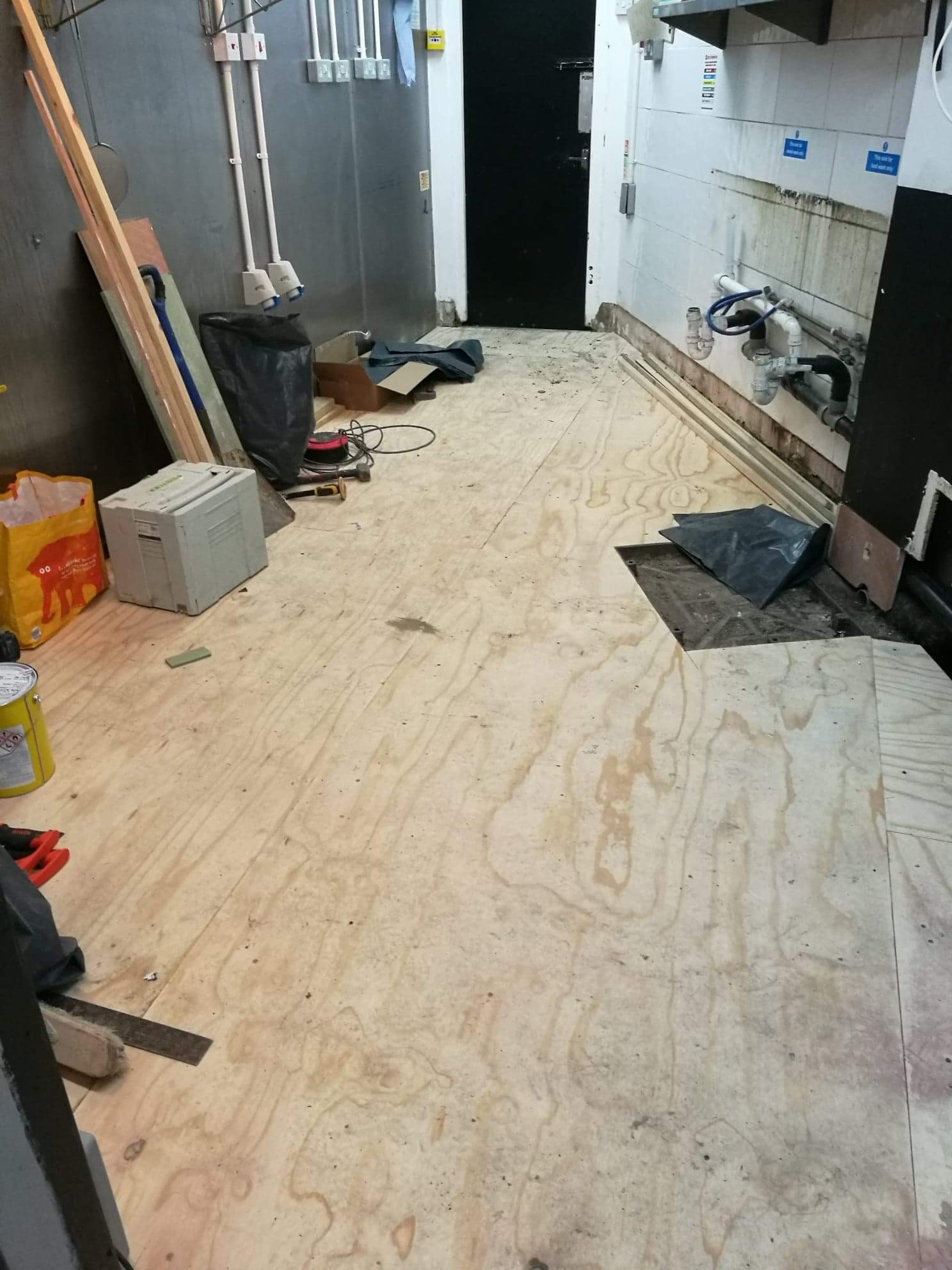 Układanie sklejki na podłodze w pomieszczeniu gospodarczym, widoczne narzędzia, worki na śmieci i niezabezpieczone rury kanalizacyjne przy ścianie.