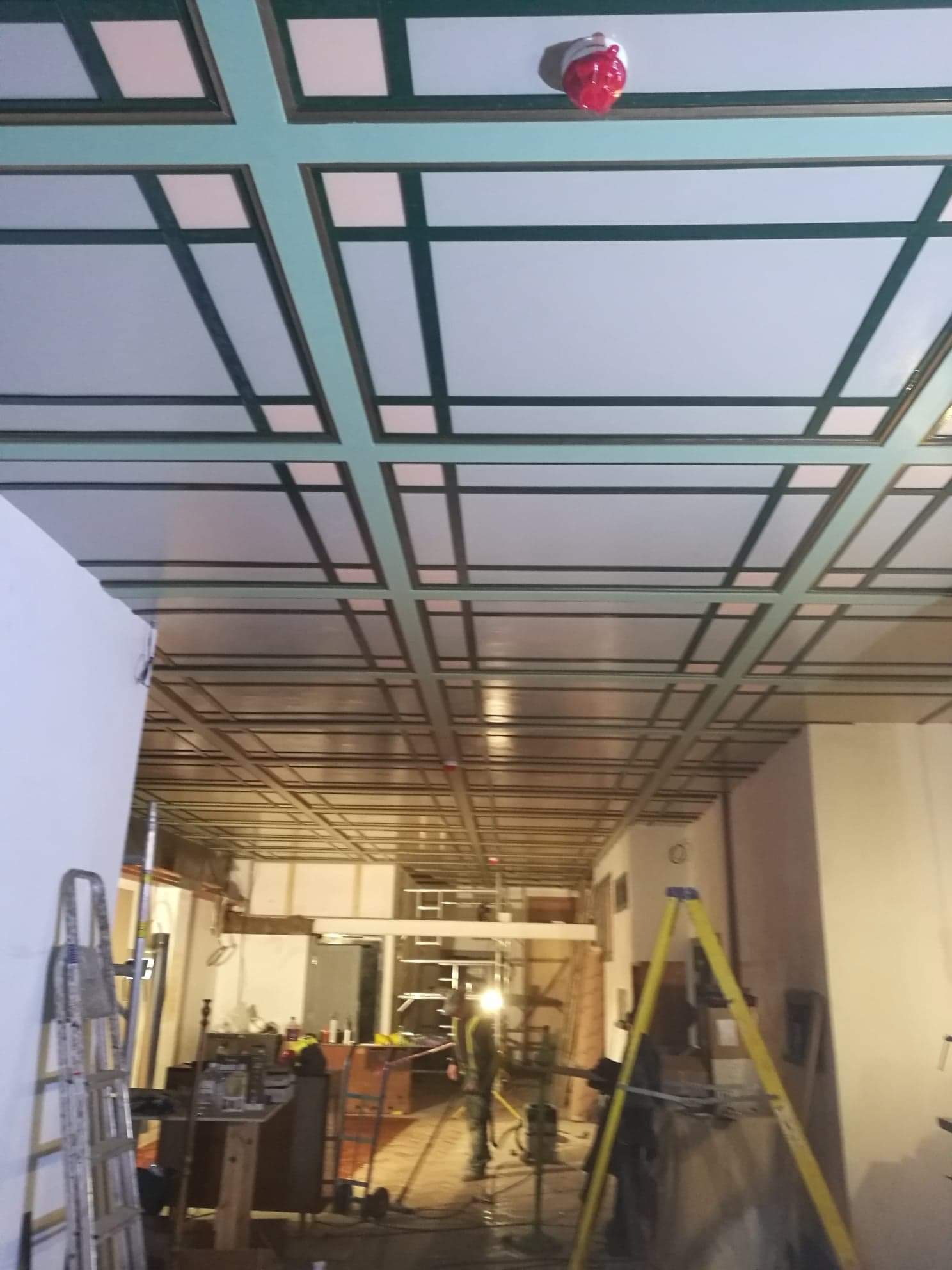 Sufit z widoczną konstrukcją nośną i częściowo zamontowanymi panelami, widoczne drabiny i sprzęt budowlany, w tle osoba w odblaskowej kamizelce.