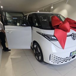 PW Kamel Jan Hadam - Biały VW ID. Buzz z czerwoną kokardą w salonie samochodowym. Otwarta boczna przesuwana szyba, widoczna część pracownika salonu. Płytki na podłodze odbijają światło.