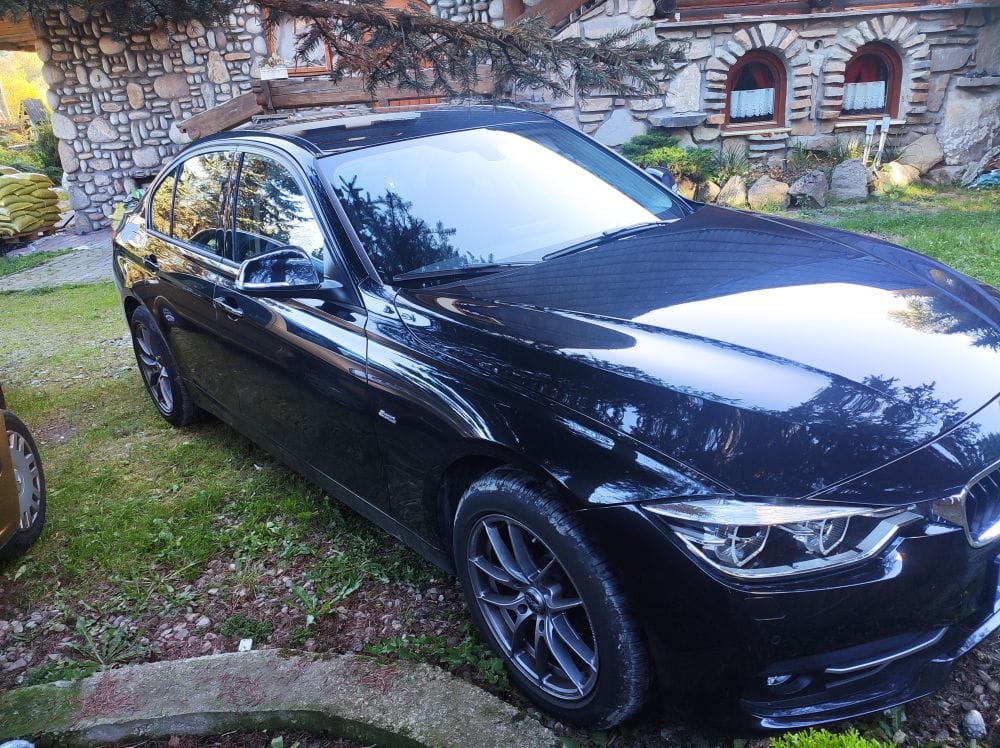 Czarny sedan BMW serii 3, zaparkowany na trawie przed kamiennym domem z ozdobnymi oknami.