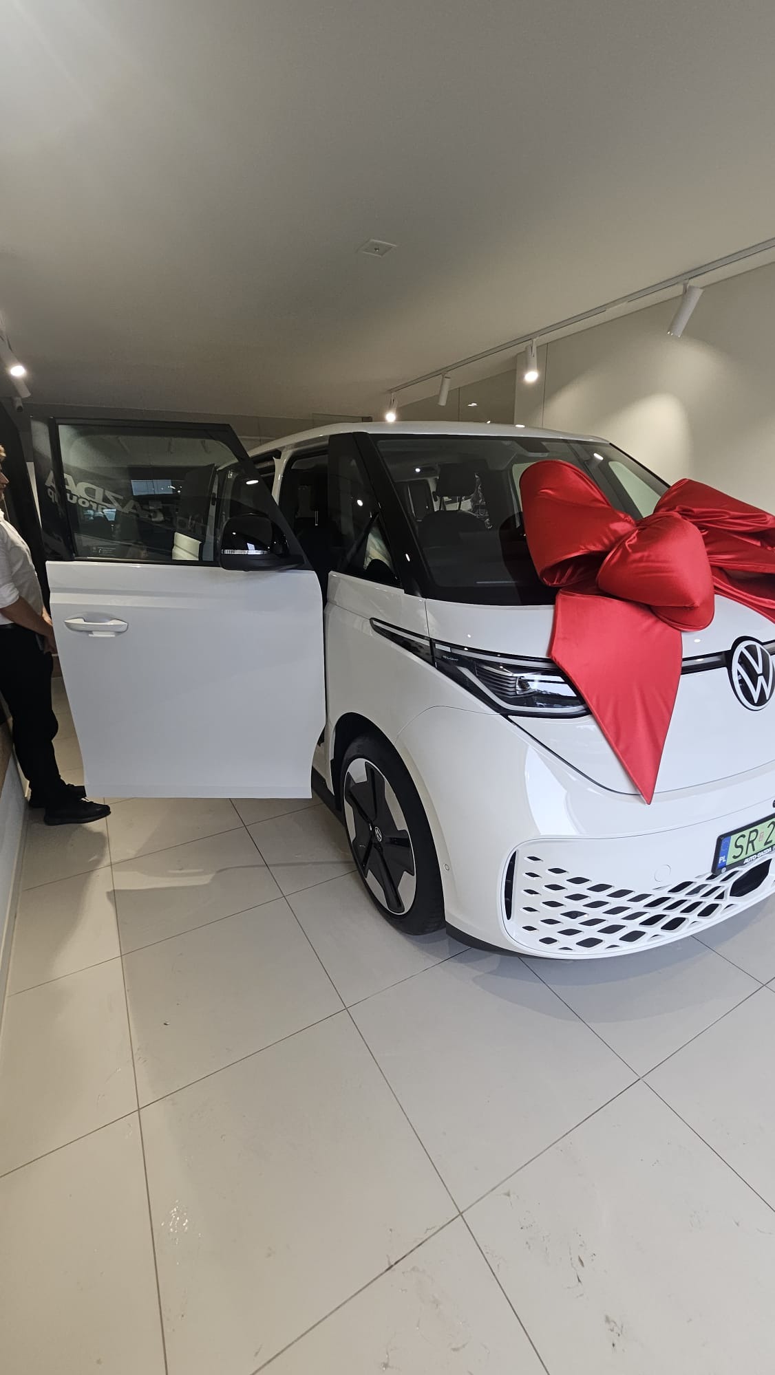 Biały VW ID. Buzz z czerwoną kokardą w salonie samochodowym. Otwarta boczna przesuwana szyba, widoczna część pracownika salonu. Płytki na podłodze odbijają światło.