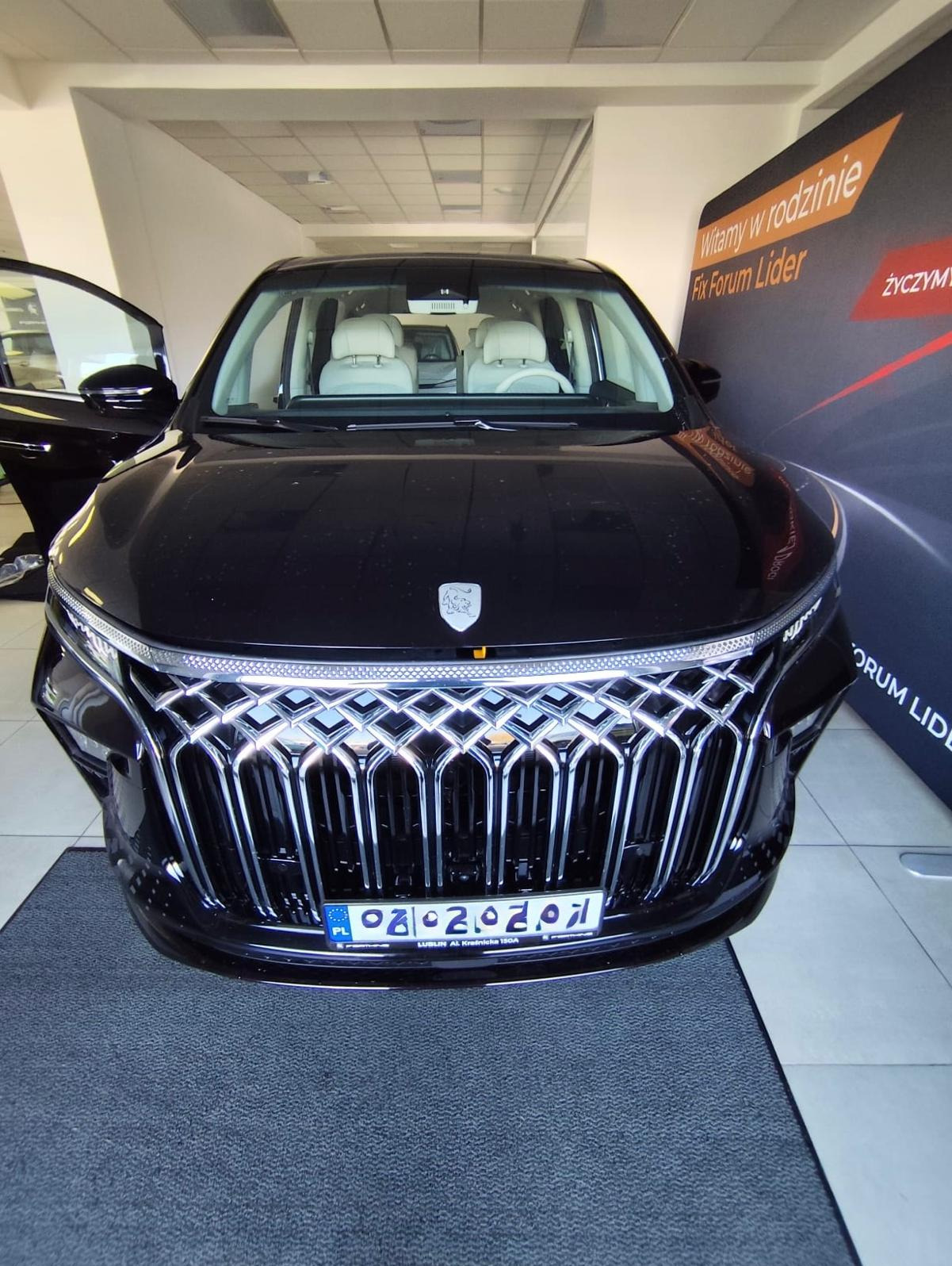 Czarny samochód osobowy z otwartymi drzwiami, widok z przodu na ozdobny grill i tablicę rejestracyjną. W tle widoczny baner reklamowy i jasne wnętrze.