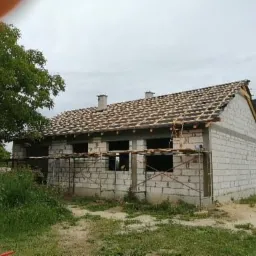 Budynek w trakcie budowy z częściowo położonym dachem z dachówki, widoczne rusztowania i ściany z białego bloczka.