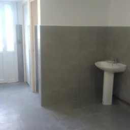 Wykończone pomieszczenie socjalne z szarą glazurą na ścianach do połowy wysokości i na podłodze, biała umywalka na postumencie, białe ściany powyżej glazury i biały sufit, widoczne fragmenty drzwi.