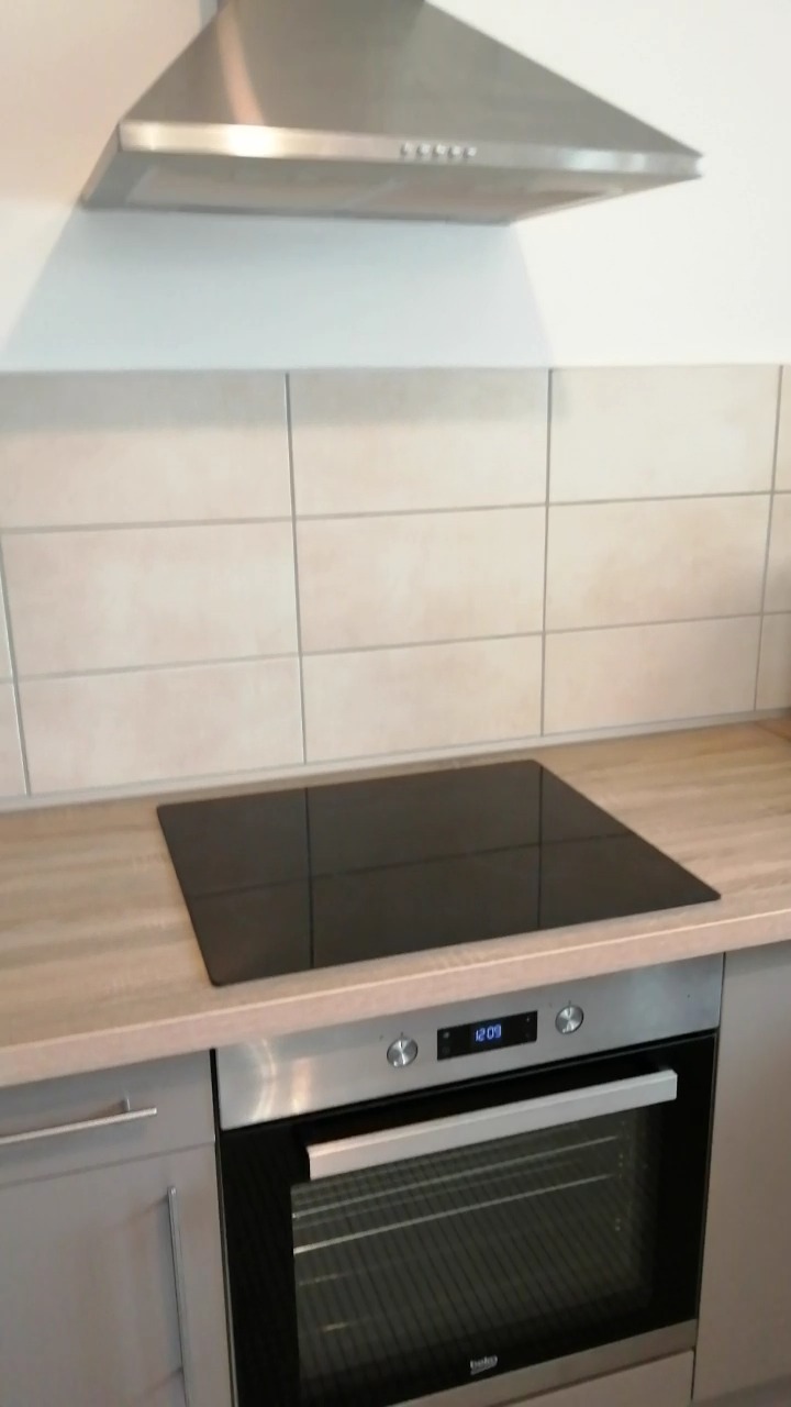 Nowoczesna kuchnia: indukcyjna płyta grzewcza na blacie w kolorze jasnego drewna, beżowe płytki na ścianie, srebrny okap i piekarnik Beko z wyświetlaczem.