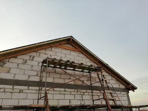 Ściana budynku w trakcie budowy z białych bloczków, widoczne rusztowanie i drewniana konstrukcja dachu pod szarym niebem.