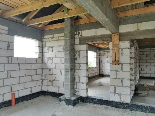 Wnętrze w trakcie budowy: ściany z białych bloczków, betonowe nadproża, drewniana konstrukcja dachu, widoczne okna i instalacja kanalizacyjna.