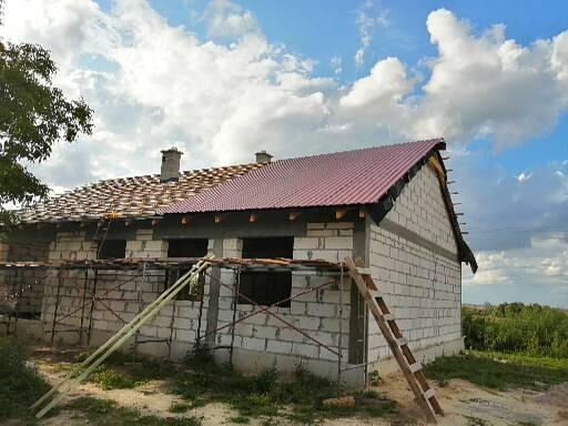 Budowa domu jednorodzinnego w trakcie: część dachu pokryta czerwoną blachą, druga część z przygotowaną konstrukcją pod dachówkę. Ściany z białego bloczka, widoczne rusztowania i drabina. Błękitne...