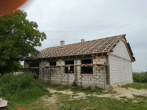 Budynek w trakcie budowy z częściowo położonym dachem z dachówki, widoczne rusztowania i ściany z białego bloczka.