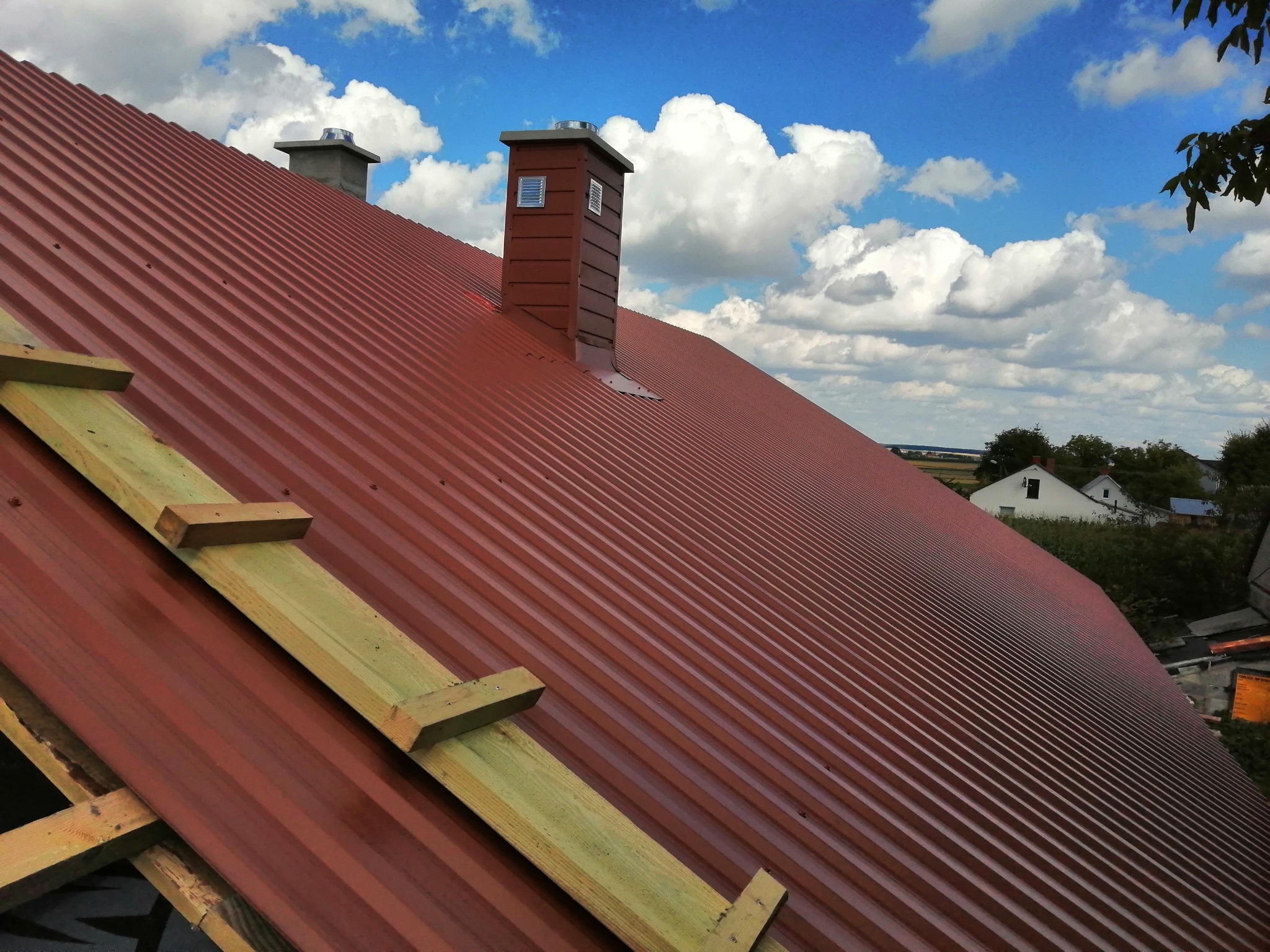 Czerwony dach z blachy trapezowej, z drewnianą konstrukcją wsporczą i kominem na tle błękitnego nieba z białymi chmurami.