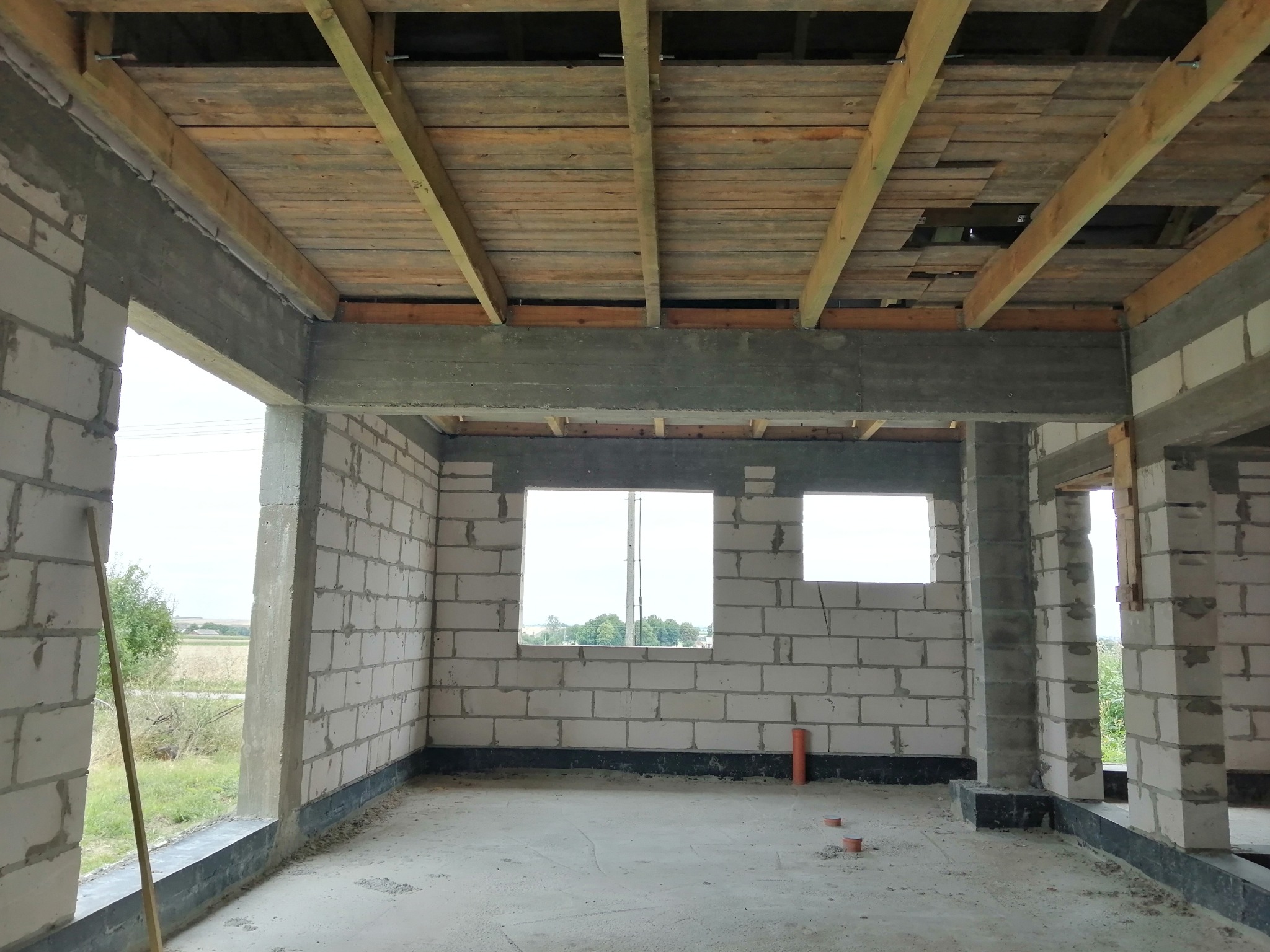 Surowy stan wnętrza budynku w trakcie budowy: ściany z bloczków, betonowe słupy i nadproża, drewniana konstrukcja stropu, widoczne okna i instalacje.