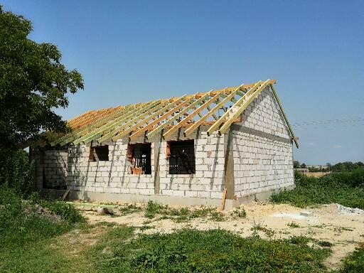 Budowa domu jednorodzinnego: ściany z bloczków betonowych, drewniana konstrukcja dachu w trakcie montażu, widoczne otwory okienne i drzwiowe.