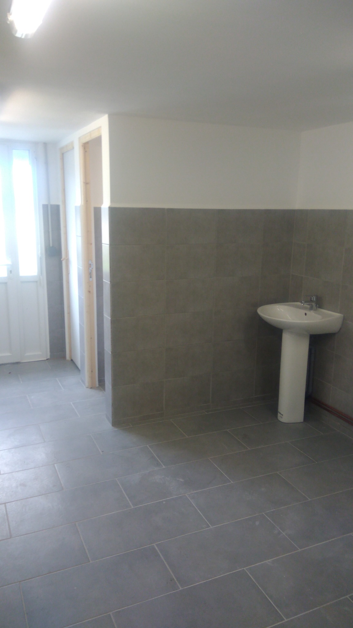 Wykończone pomieszczenie socjalne z szarą glazurą na ścianach do połowy wysokości i na podłodze, biała umywalka na postumencie, białe ściany powyżej glazury i biały sufit, widoczne fragmenty drzwi.