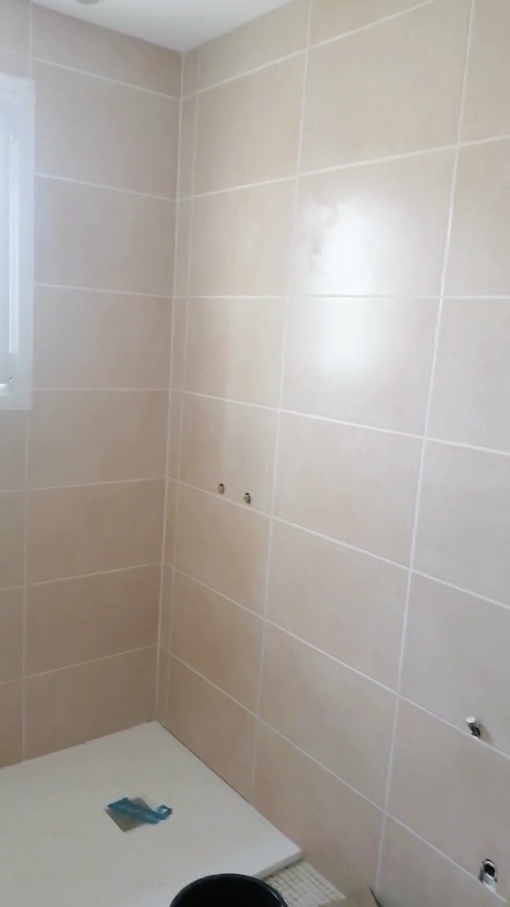 Ściana łazienki wyłożona beżowymi płytkami ceramicznymi z widocznymi otworami na armaturę, biały brodzik i czarne wiadro na podłodze.