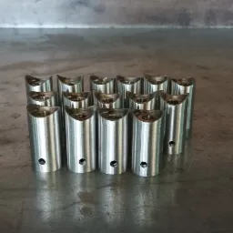 Szereg metalowych, cylindrycznych elementów z wycięciem w kształcie półksiężyca i otworem bocznym, ułożonych na szarej, metalowej powierzchni.