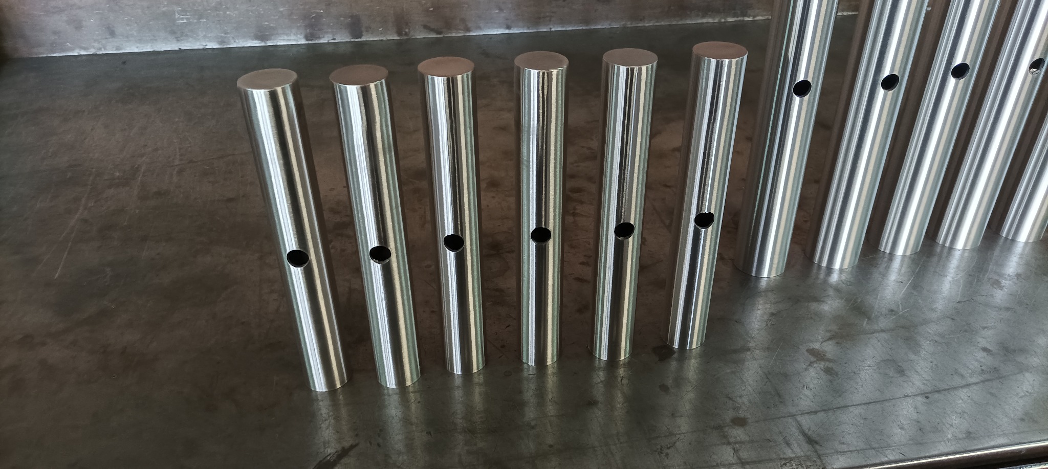 Rząd polerowanych, cylindrycznych elementów metalowych z otworem, ułożonych na metalowym stole warsztatowym, widok z góry pod kątem.