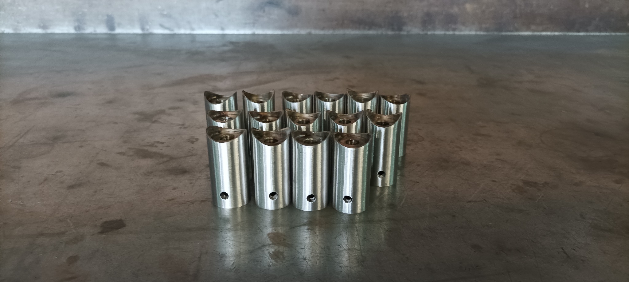Szereg metalowych, cylindrycznych elementów z wycięciem w kształcie półksiężyca i otworem bocznym, ułożonych na szarej, metalowej powierzchni.