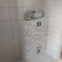 Obudowa stelaża podtynkowego WC wykończona płytkami ceramicznymi z dekoracyjnym wzorem, widoczne przyłącze kanalizacyjne i skrzynka rewizyjna, obok ściana z jasnych płytek.