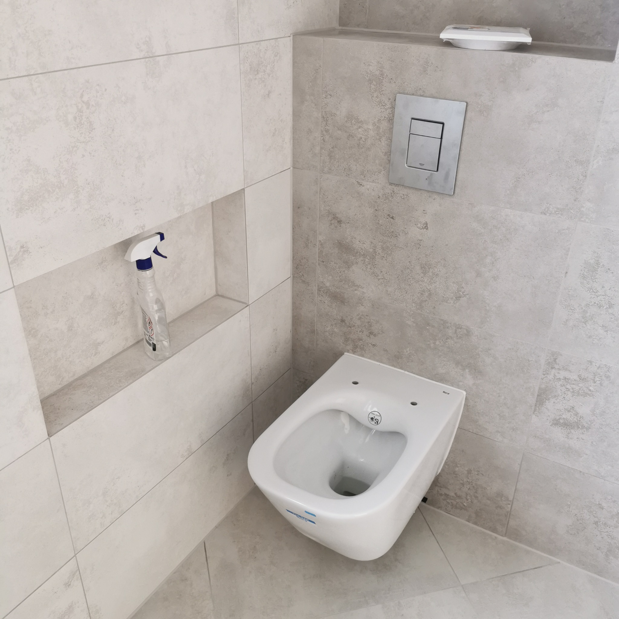 Wisząca toaleta z podtynkową spłuczką Grohe na ścianie wyłożonej szarymi płytkami, obok wnęka z butelką płynu do czyszczenia, widok na fragment podłogi z płytek.