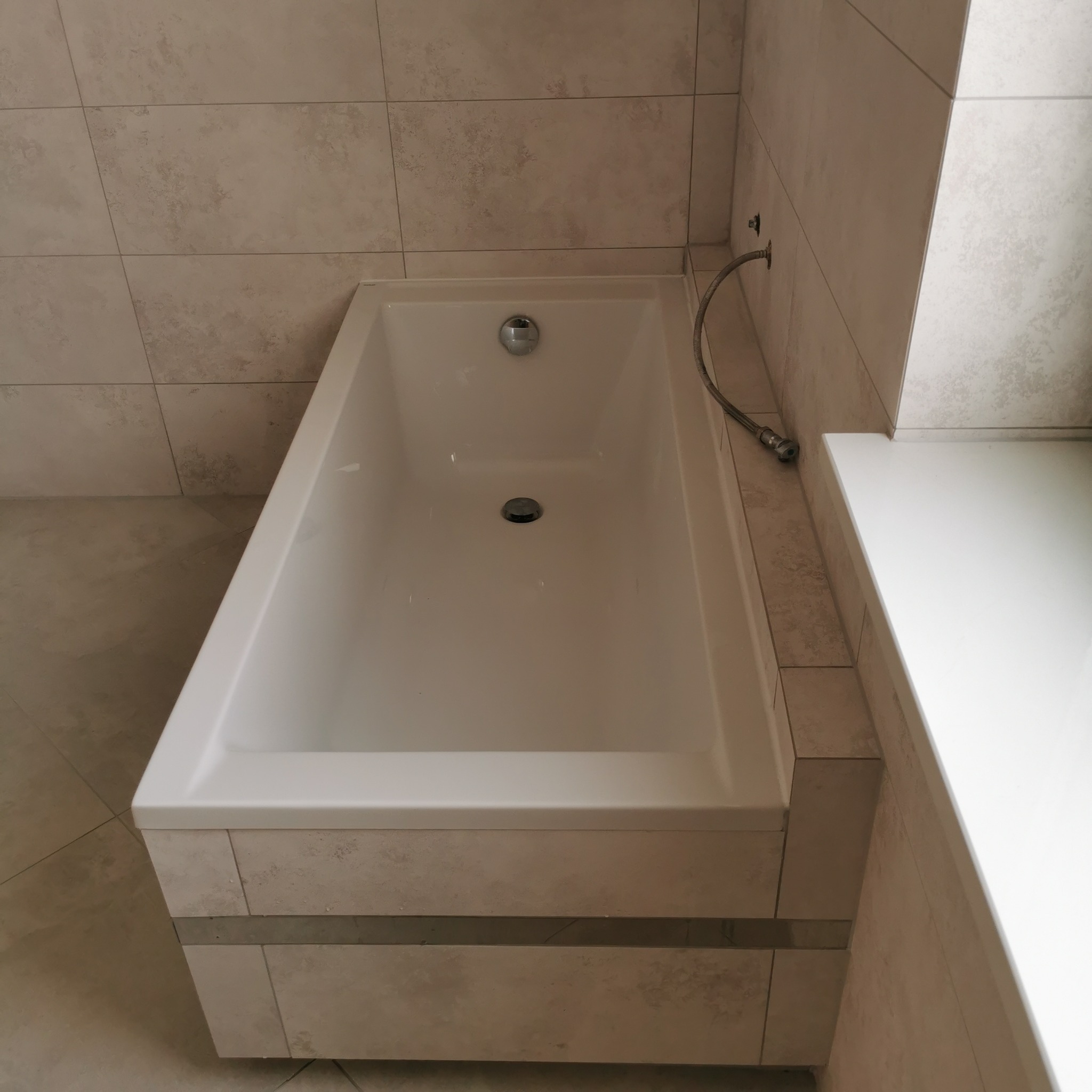 Biała, prostokątna wanna obudowana płytkami imitującymi kamień, z chromowaną armaturą i odpływem, na tle ściany wyłożonej identycznymi płytkami.