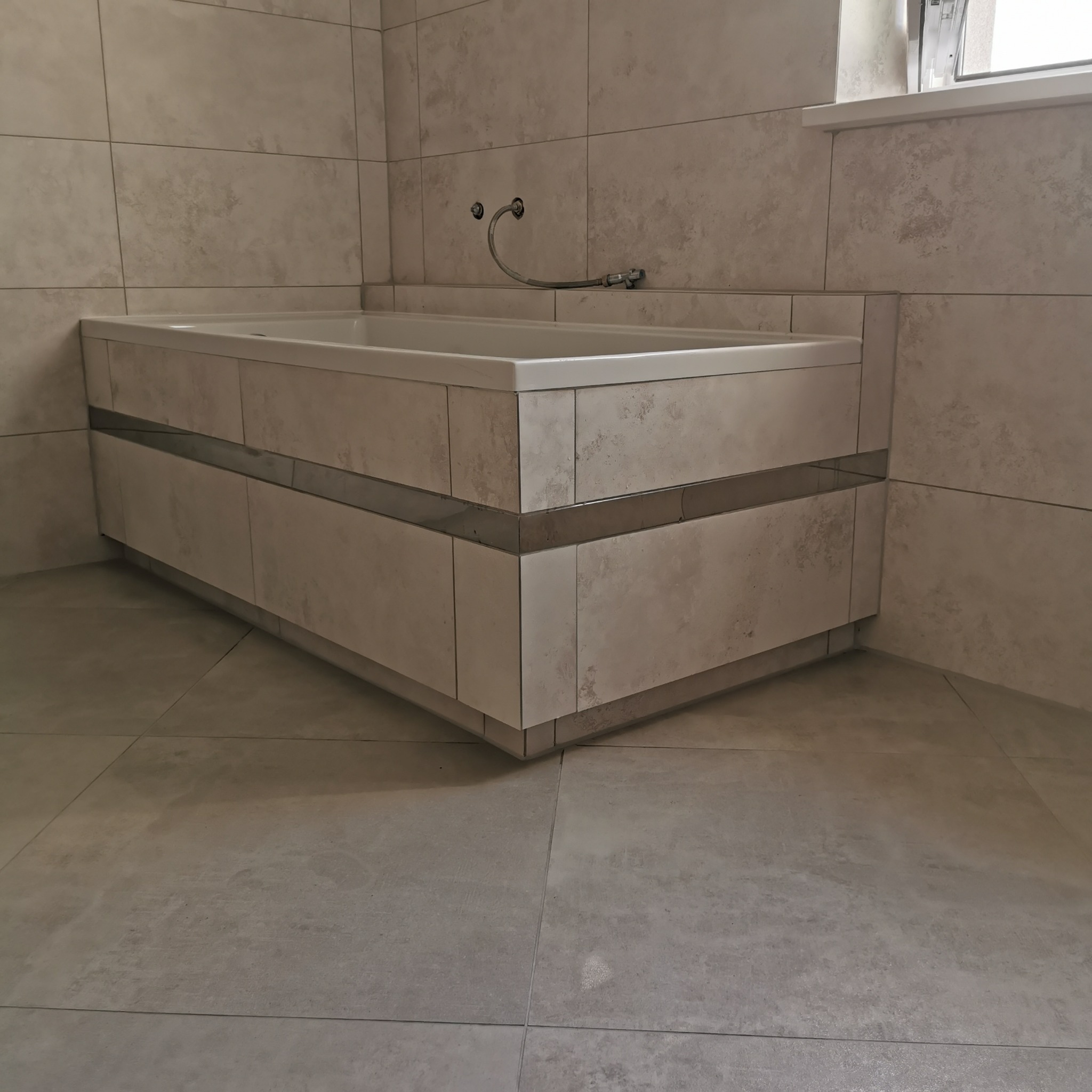 Obudowana wanna z jasnymi płytkami o wzorze betonu, widoczne przyłącza wodne, wykończenie listwą dekoracyjną, podłoga z płytek w podobnym odcieniu.