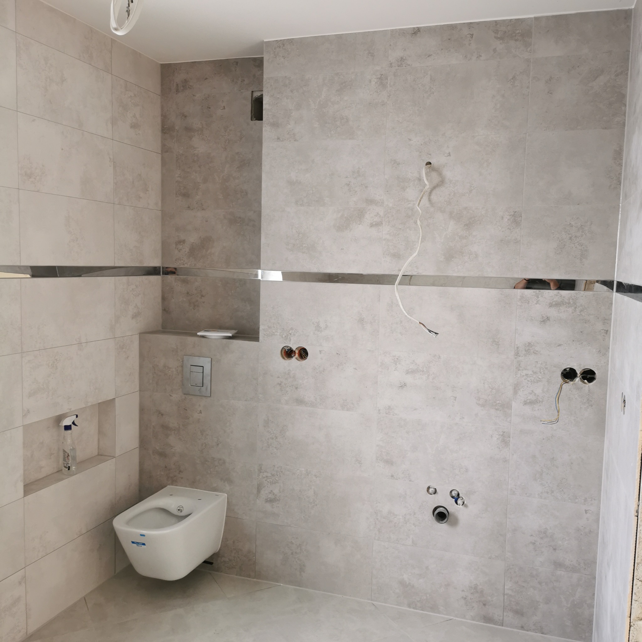 Łazienka w trakcie remontu: szare płytki z metalową listwą, wiszący bidet, przygotowane punkty elektryczne i hydrauliczne, widoczne przewody.