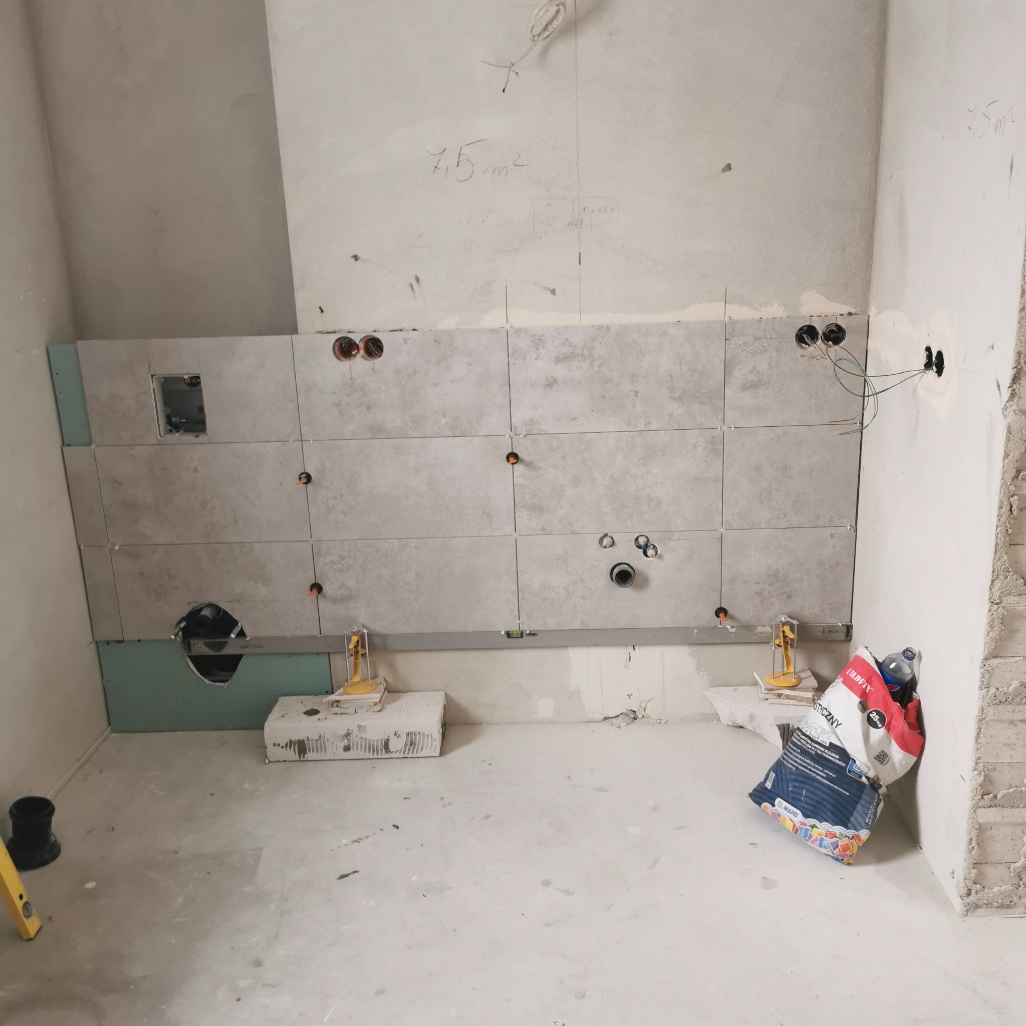 Ściana w trakcie układania płytek w łazience w Ostrowie Wielkopolskim, widoczne narzędzia glazurnicze, poziomica, paczka kleju do płytek oraz wywiercone otwory pod instalacje.