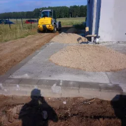 Przygotowanie podłoża pod budowę: ż&oacute;łta ładowarka Kramer Allrad na tle pola, pryzmy piasku i fragment budynku w stanie surowym, cień osoby w dolnej części kadru.