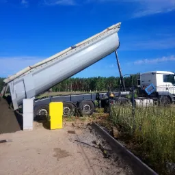Wyładowany piasek z ciężarówki na placu budowy, widoczne obrzeża chodnika, słupki ogrodzeniowe i zwinięty przedłużacz elektryczny.