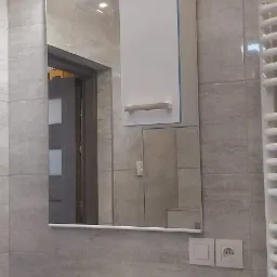 Nowoczesna łazienka z szarymi płytkami, białym umywalką i szafką z lustrem oraz oświetleniem LED. W tle widoczne drzwi.