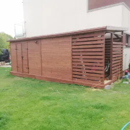 Drewniana konstrukcja z zamykaną częścią i otwartą przestrzenią z poziomych desek, prawdopodobnie schowek na narzędzia lub sprzęt ogrodowy, ustawiona na trawniku obok budynku mieszkalnego.