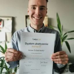 Uśmiechnięty mężczyzna prezentuje dyplom ukończenia kursu IT Python Developer od Coders Lab, trzymając go oburącz na tle jasnej ściany z roślinami doniczkowymi.