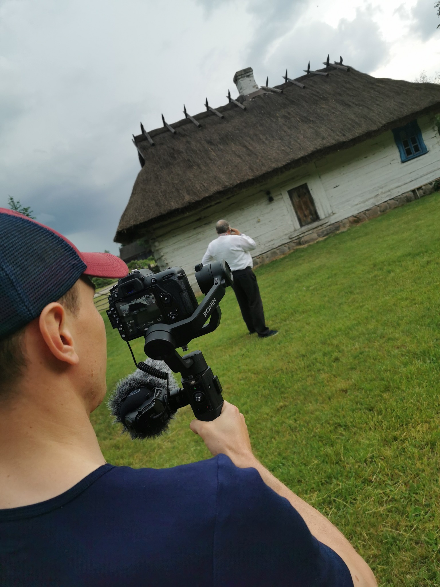 Operator z gimbalem i mikrofonem filmuje starą drewnianą chatę z krytym strzechą dachem na tle zielonego trawnika i pochmurnego nieba.