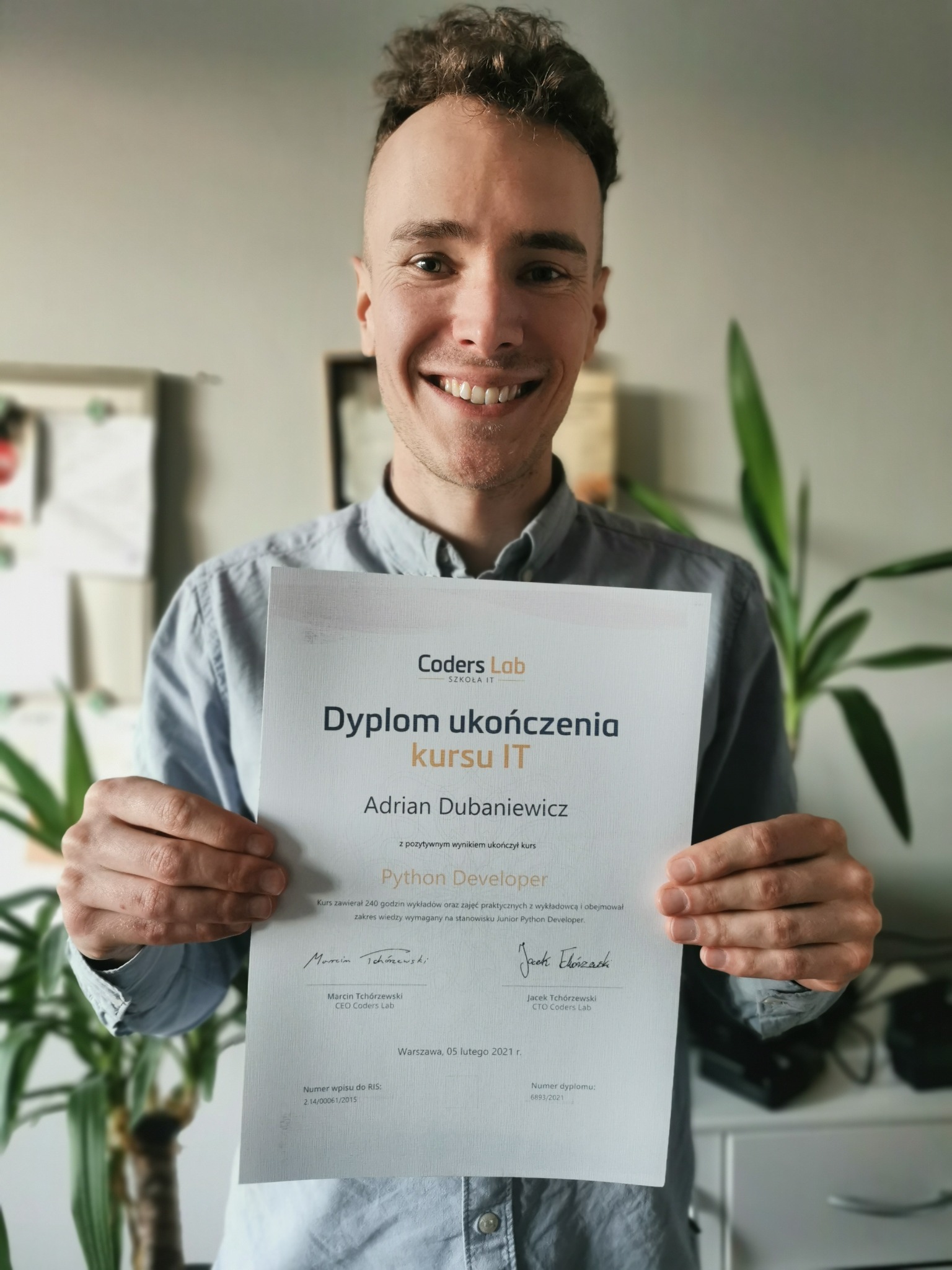 Uśmiechnięty mężczyzna prezentuje dyplom ukończenia kursu IT Python Developer od Coders Lab, trzymając go oburącz na tle jasnej ściany z roślinami doniczkowymi.