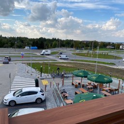 Z-K Ogród Zbigniew Nędza - Widok z góry na nowo zagospodarowany teren przydrożny z parkingiem, chodnikiem z kostki brukowej, pasem zieleni i miejscami do siedzenia pod parasolami McDonald's, widoczny ruch samochodowy...
