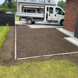 Przygotowany teren pod trawnik z rolki, widoczne obrzeża i siatka wzmacniająca, w tle budynek mieszkalny i samochód dostawczy