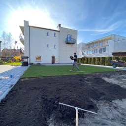 Z-K Ogród Zbigniew Nędza - Układanie trawy z rolki na przygotowanym gruncie przy nowoczesnym domu, widoczny pracownik z rolką trawy, grabie i częściowo ułożona trawa, słoneczny dzień.