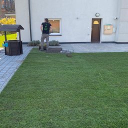 Z-K Ogród Zbigniew Nędza - Układanie trawy z rolki przy nowym domu z brązową studnią na posesji i pracownikiem w koszulce z logo firmy ogrodniczej.