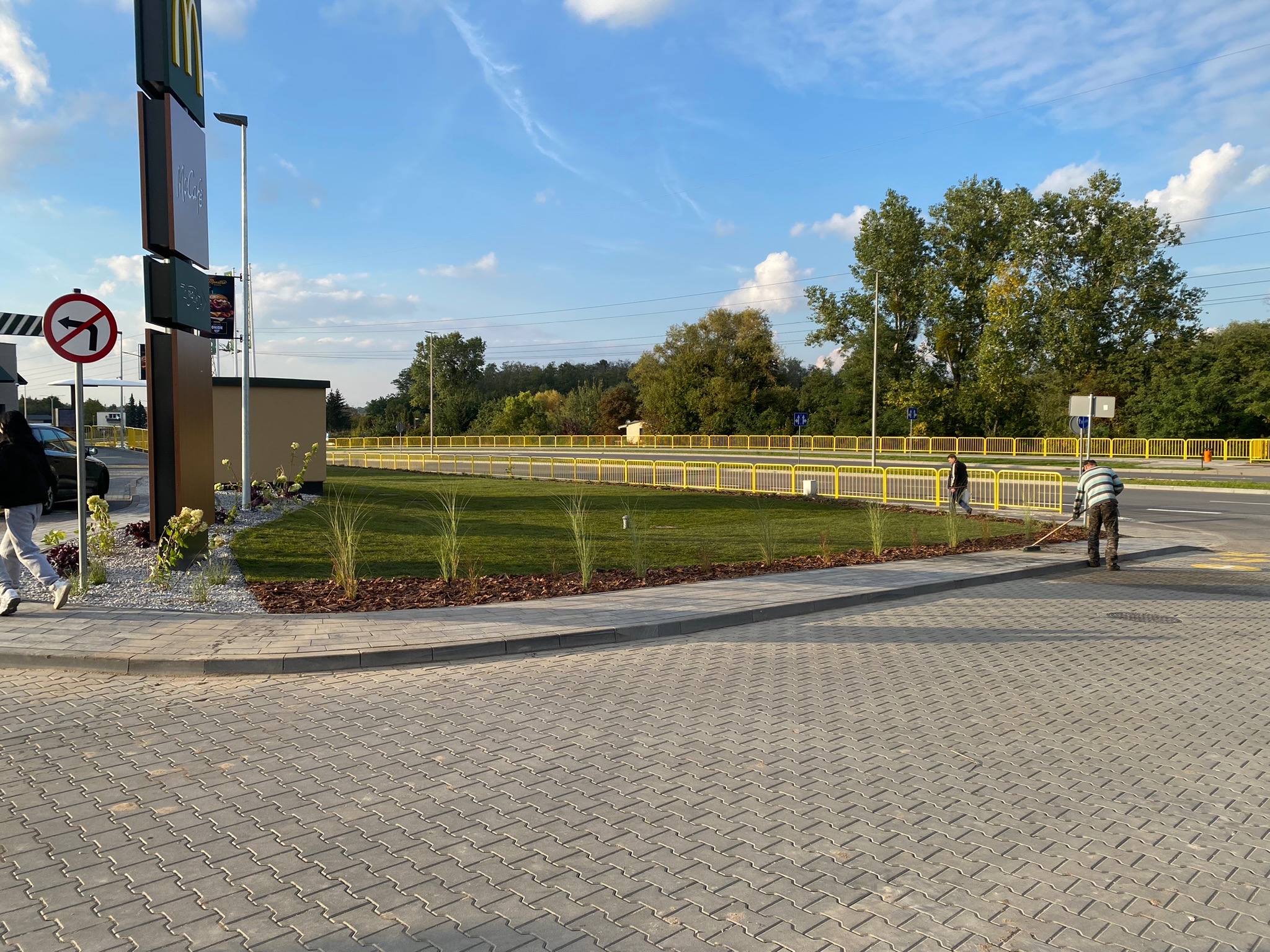 Pracownik w paski zamiata chodnik z kostki brukowej przy nowo założonym trawniku z ozdobnymi trawami i korą, w tle żółte barierki i znak McDonalda.