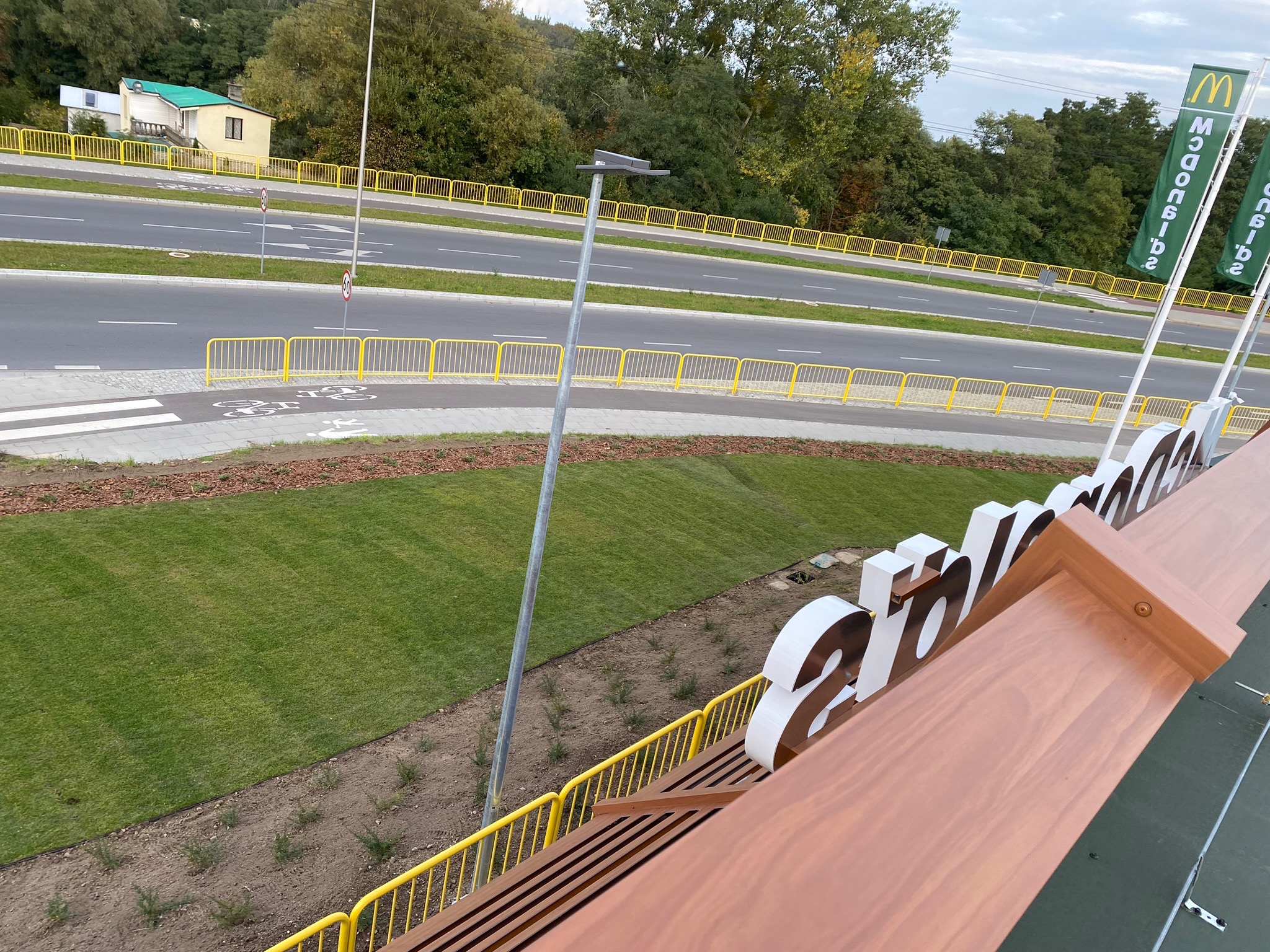 Widok z góry na świeżo założony trawnik z rolki przy drodze, z widocznymi elementami infrastruktury drogowej i budynkami w tle.